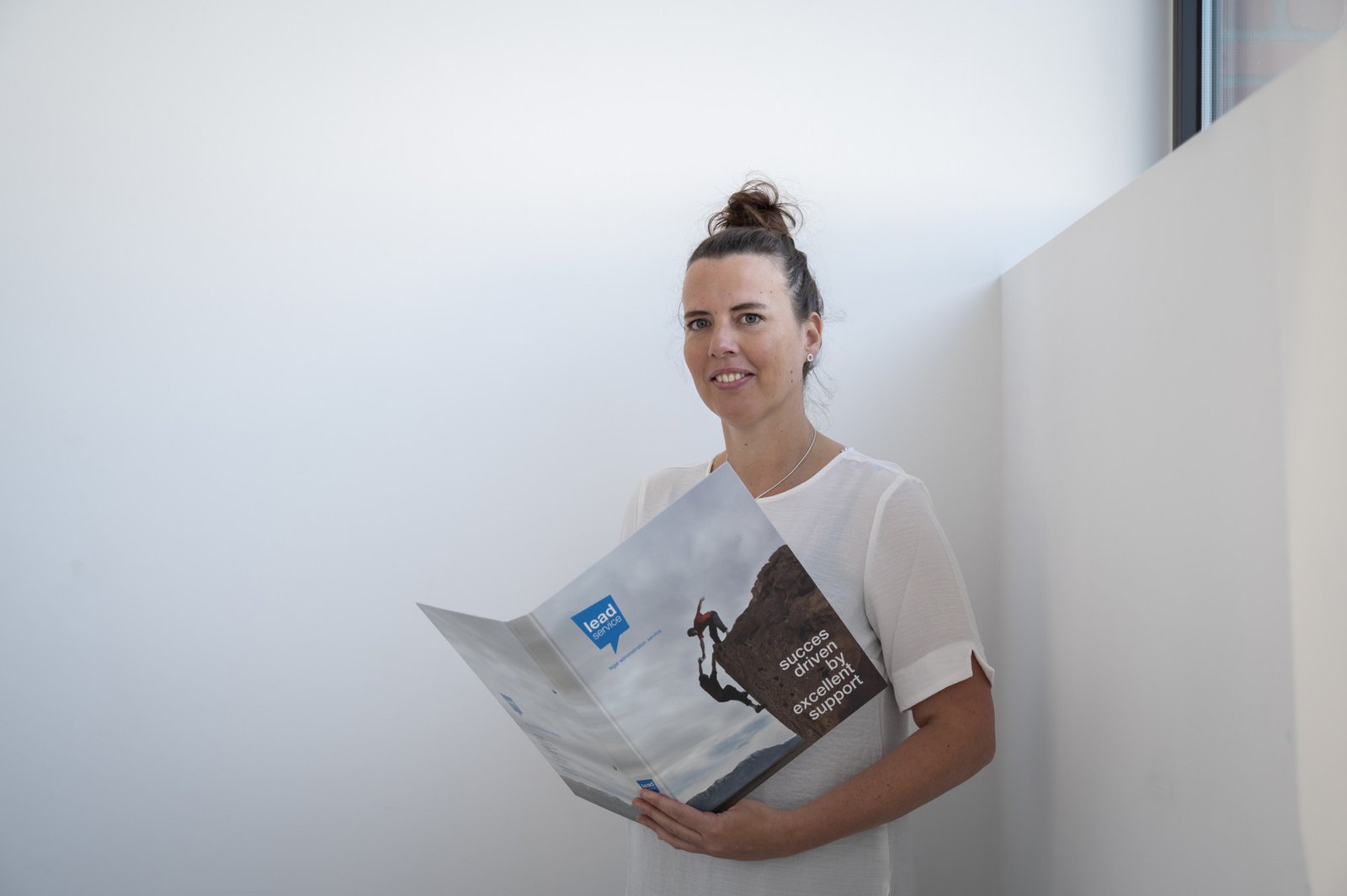 Een vrouw die een brochure of tijdschrift over "team bedrijf oprichten" vasthoudt terwijl ze binnen staat en met een lichte glimlach buiten de camera kijkt.
