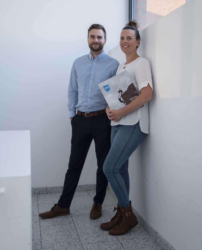 Een man en een vrouw staan in een lichte hoek; de vrouw houdt een map vast, ze zijn allebei casual gekleed en kijken naar de camera.